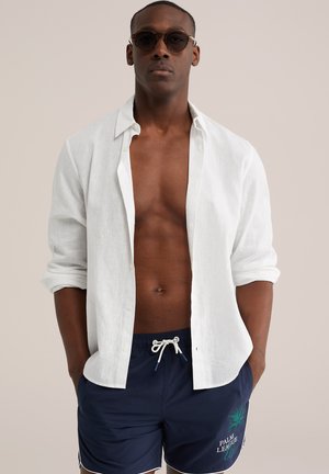 Mężczyzna w okularach przeciwsłonecznych, rozpiętej białej koszuli z podwiniętymi rękawami oraz granatowych kąpielówkach z troczkiem i logo palmy, z rękami w kieszeniach.