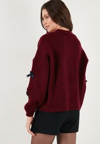 Burgundyröd stickad tröja med ribbad textur, oversized passform och svarta banddetaljer på ärmarna. Enkelt, avslappnat design.