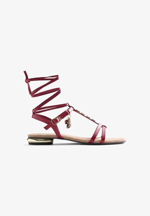 Sandale plate bordeaux avec brides chevillées, boucle dorée, petits charms et talon bas avec accents métalliques.