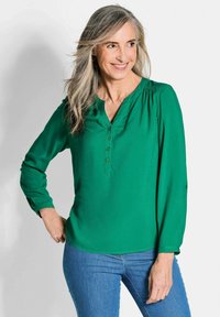 Blusa verde a maniche lunghe con scollatura a V e dettagli con bottoni. Realizzata in tessuto morbido, presenta arricciature sulle spalle. Abbinata a jeans blu.