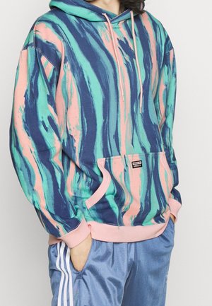 Osoba nosząca wielobarwną bluzę z kapturem typu tie-dye w kolorach niebieskim, zielonym i różowym z różowymi mankietami, w zestawie z niebieskimi spodniami dresowymi Adidas z białymi paskami.