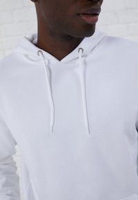 Sudadera blanca con capucha ajustable, ojales plateados y un acabado de tela suave y texturizada. Vista en primer plano que muestra el área del cuello y el detalle de la tela.