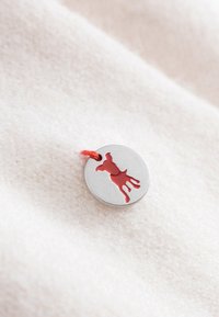 Metalowa zawieszka z wyciętym konturem czerwonego psa, przymocowana cienką czerwoną nitką, leżąca na strukturalnej, jasnobeżowej tkaninie.