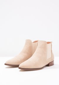 Botas de tobillo de ante beige con paneles elásticos en los laterales, punta afilada y costuras decorativas blancas en el empeine. Tacón de madera plano.