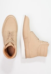 Beżowe skórzane buty typu high-top o gładkiej fakturze, zaokrąglonym nosku, płaskiej białej podeszwie i systemie sznurowania. Posiadają przeszywane detale.