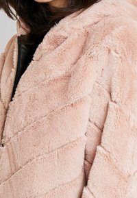 Chaqueta de pelaje sintético rosa claro con patrón de flechas, ajuste oversize y detalle de cremallera negra. Textura suave y material esponjoso.