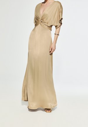 Femme portant une longue robe beige soyeuse avec un décolleté en V profond et un détail torsadé à l’avant, debout devant un fond clair uni.