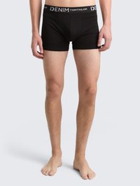 Manlig modell som bär svarta jeansboxershorts från Tom Tailor, står barfota mot en enkel ljus bakgrund.