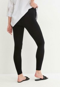 Leggings noirs avec une coupe slim, fabriqués en tissu extensible. Portés avec une chemise blanche, montrant une texture lisse et une finition sans couture.