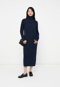 Nainen seisoo päällään tummansininen palmikkoneuleinen poolopuku, mustat loafer-kengät ja musta olkahihnallinen laukku, taustalla valkoinen seinä.
