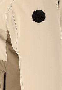 Beige buitenjack met een zachte textuur, een contrasterend bruin paneel en een zwart circulair logo dat is geperst met een bergontwerp.