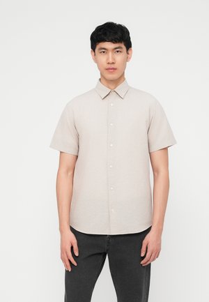 Νεαρός άνδρας όρθιος, φοράει ανοιχτό μπεζ κοντομάνικο πουκάμισο με κουμπιά και σκούρο γκρι παντελόνι, με φόντο απλό λευκό.