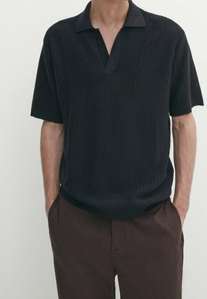 Homme portant un polo noir texturé à manches courtes avec col en V et pantalon marron foncé, mains dans les poches.