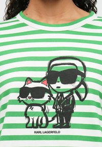 Zöld és fehér csíkos póló, fekete-fehér mesefigura nyomattal, ahol egy karakter és egy napszemüveges macska látható, "KARL LAGERFELD" felirattal.