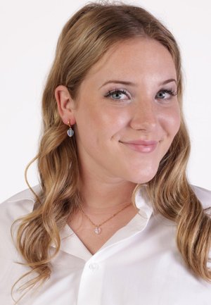 Gold hoop earrings with a blue heart-shaped accent. The model wears a white button-up shirt and a gold chain necklace with a heart pendant.