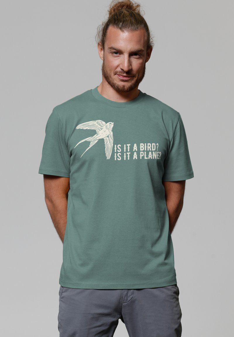 Grünes Baumwoll-T-Shirt mit einem Vogeldruck und dem Text "Ist es ein Vogel? Ist es ein Flugzeug?" in cremefarbener Schrift. Kurze Ärmel und Rundhalsausschnitt.