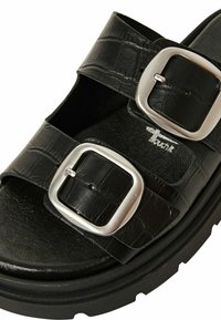 Svart sandal tillverkad av texturerat syntetmaterial, med två breda remmar, silverfärgade metallspännen och en tjock sula med ett mönster.