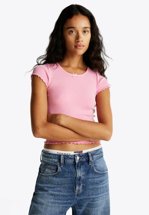 Top corto rosa a costine con maniche corte, caratterizzato da bordi scallop e un piccolo fiocco decorativo al collo, abbinato a jeans blu.