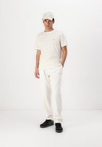 Gaiši krēma t-krekls ar smalku rakstu, apģērbts ar baltām biksēm un melniem čības. Modelis valkā beidzīgu cepuri, stāvot uz balta fona.