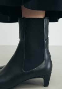 Schwarzer Knöchelstiefel mit einer glatten Lederoberfläche, elastischen Seitenteilen und einem niedrigen, verjüngten Absatz. Das Design zeichnet sich durch eine elegante Silhouette aus.