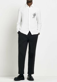 Camicia bianca a maniche lunghe con stampato floreale nero sulla parte sinistra del petto, abbinata a pantaloni neri e scarpe nere, presentando un design pulito.