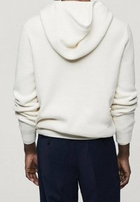 Persona che indossa un hoodie lavorato a maglia bianco con maniche rilassate e pantaloni blu scuro, mostrata di spalle contro uno sfondo chiaro e semplice.
