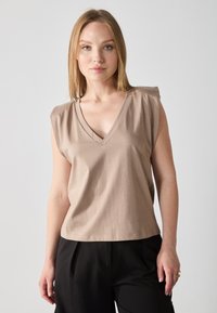 Top sin mangas en color beige con escote en V, confeccionado en una tela suave. Presenta sutiles pliegues en los hombros y un corte holgado, combinado con pantalones negros.
