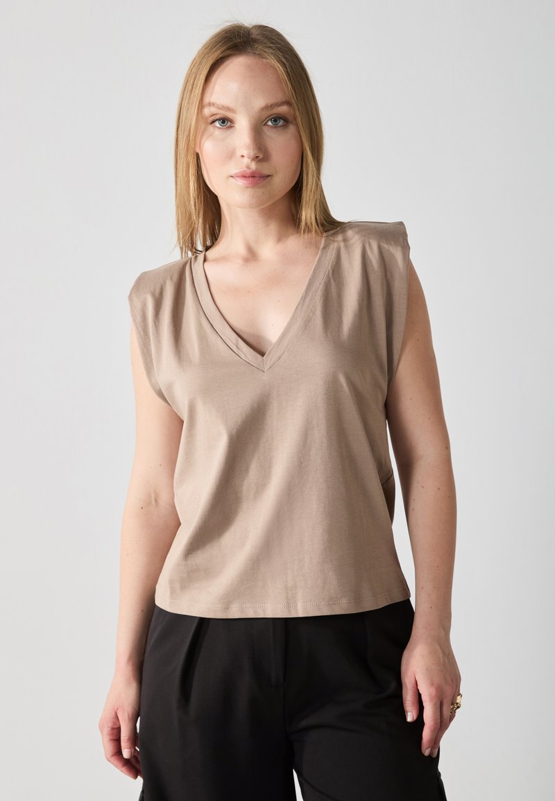 Top sin mangas en color beige con escote en V, confeccionado en una tela suave. Presenta sutiles pliegues en los hombros y un corte holgado, combinado con pantalones negros.