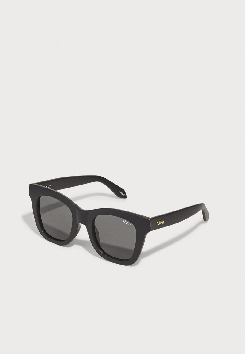 Lunettes de soleil rectangulaires noires avec une finition mate, dotées de verres sombres et d'un logo doré subtil sur le côté. Monture épaisse et audacieuse.