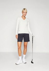 Γυναίκα με κοντά ξανθά μαλλιά, ντυμένη με λευκή ζακέτα, μαύρες σορτς, άσπρα αθλητικά παπούτσια και γα gloves, κρατώντας μια ρακέτα γκολφ, στέκεται σε απλό φόντο.