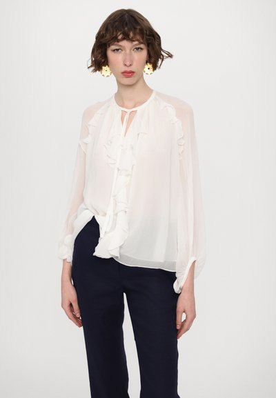 White, semi-sheer blouse with ruffled details down the front and around the neckline. Long, billowy sleeves and a relaxed fit. Paired with dark trousers.