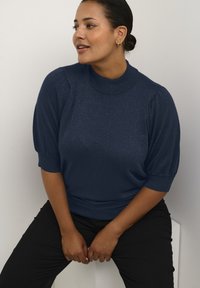 Maglione blu navy a maniche corte con colletto alto e finitura testurizzata. Presenta maniche a sbuffo e una vita aderente. Pantaloni neri indossati sotto.