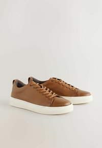 Zapatillas de cuero marrón con un acabado texturizado, que cuentan con una suela de goma blanca, punta redondeada y un diseño clásico de cordones.