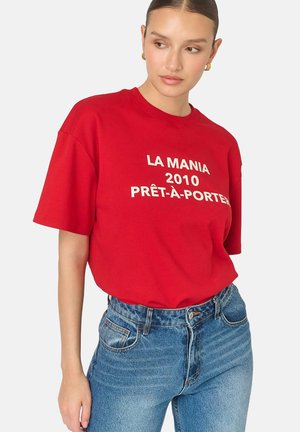 Červené bavlnené voľné tričko s bielym textom "LA MANIA 2010 PRÊT-À-PORTER," skombinované s modrými nohavicami s vysokým pásom.