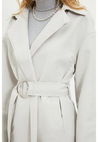 Mujer vistiendo un abrigo trench de color beige claro con cinturón sobre un suéter de cuello alto gris, con una cadena de plata visible.