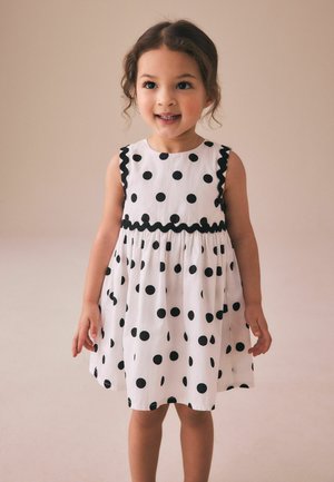 Jeune fille souriante aux cheveux bouclés portant une robe blanche sans manches à pois noirs avec bordure noire, debout devant un fond uni.