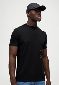 Μαύρο βαμβακερό μπλουζάκι με κοντά μανίκια, στρογγυλή λαιμόκοψη και μπροστινή τσέπη που διαθέτει χρυσό σχέδιο λογότυπου. Το μοντέλο φοράει ένα σκούρο καπέλο.