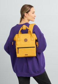 Yellow backpack with a square shape, featuring a top handle, adjustable strap, front zip pocket, and a round logo patch. Textured fabric.