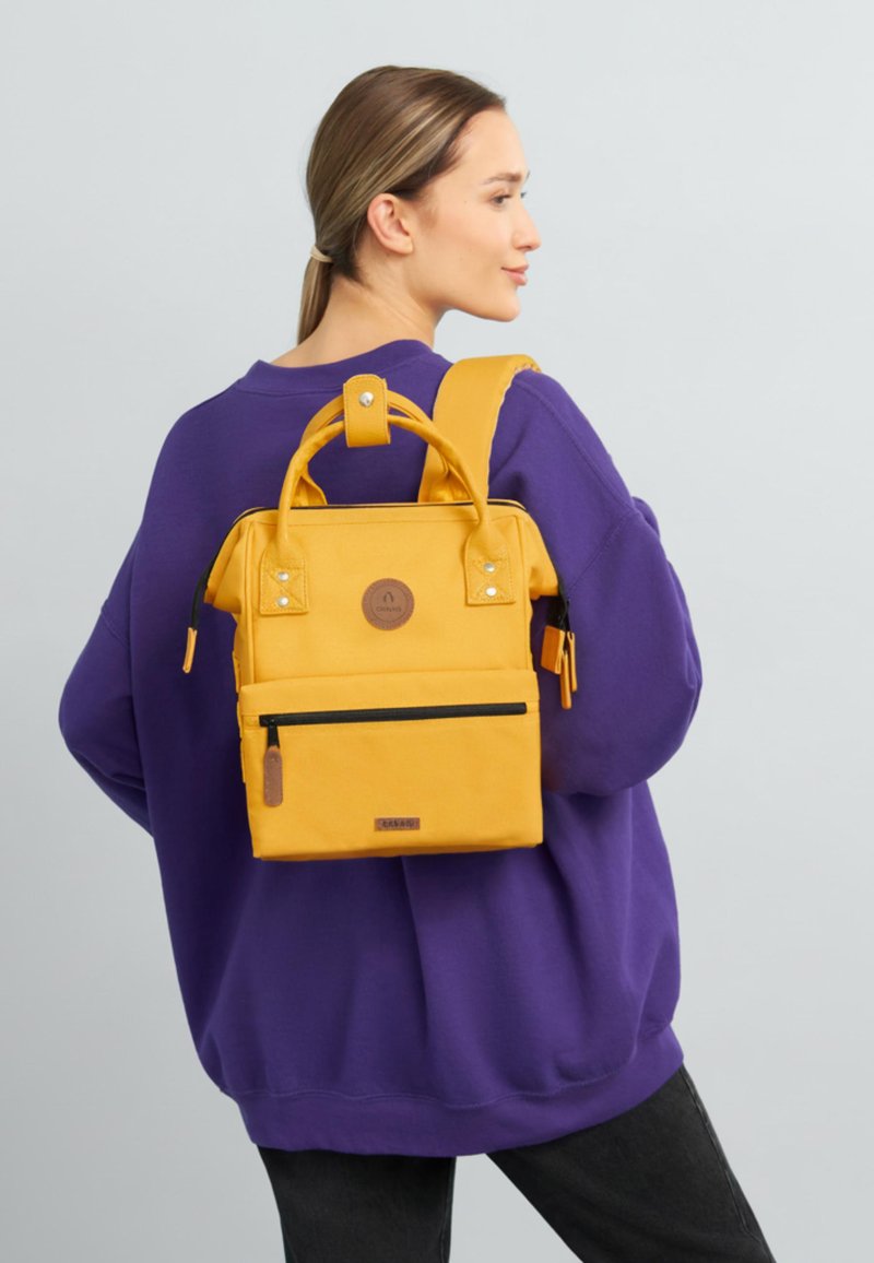 Yellow backpack with a square shape, featuring a top handle, adjustable strap, front zip pocket, and a round logo patch. Textured fabric.