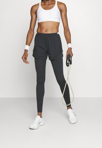 Pantalones cortos deportivos grises sobre mallas negras, combinados con un sujetador deportivo blanco y zapatillas deportivas blancas. En una mano sostiene una banda de resistencia.