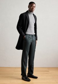 Manteau noir long sur un pull à col roulé gris, associé à un pantalon plissé vert et des chaussures habillées noires. Textures lisses et coupe ajustée.