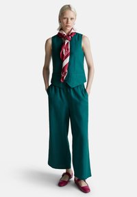 Donna bionda che indossa un gilet senza maniche color turchese e pantaloni larghi, sciarpa a strisce rosse e bianche, e scarpe basse rosse, in piedi con le mani nelle tasche.