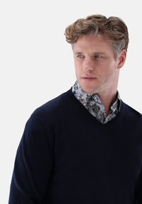 Navy V-Ausschnitt-Pullover aus weichem Material, über einem hellen, blumigen Hemd getragen. Mit Knöpfen und einem detaillierten Blumenmuster am Kragen.
