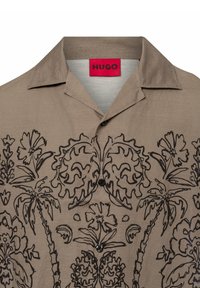 Bruine shirt met zwarte bloem- en palmbladontwerpen, korte kraag en knoopsluiting. Etiket leest "HUGO" in het rood. Gladde textuur.