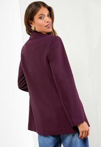 Bordeaux blazer med skræddersyet pasform, lange ærmer og strukturerede skuldre. Glat tekstur, parret med blå denimjeans. Guldøreringe synlige.