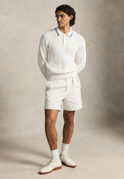 Homme debout portant un pull en maille torsadée blanche, un short plissé blanc, des chaussettes blanches et des chaussures habillées blanches sur un fond neutre.