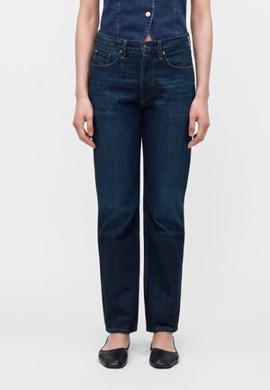 Personne portant un jean droit bleu foncé, un top bleu sans manches avec un bouton, et des chaussures plates noires, debout devant un fond uni.
