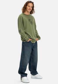 Man with curly hair wearing an olive green long-sleeve shirt with graphics, loose dark jeans, and white sneakers, standing with hands in pockets.