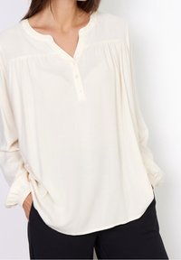 Chemisier en crème clair avec un décolleté en V, des détails plissés et des manches longues. Tissu doux avec une coupe décontractée. Associé à un pantalon noir.