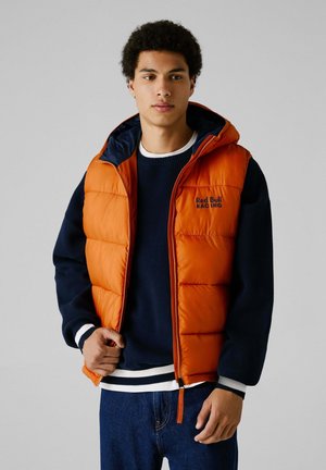 Vaddern orange väst med huva, med marinblå foder och logotyp. Bärs över en marinblå tröja med vita ränder vid ärmarna och fållen. Jeansbyxor.